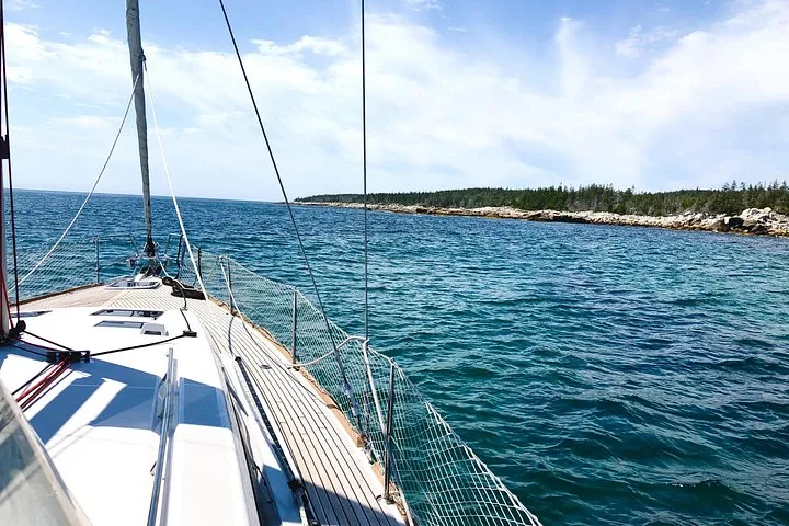 Vue par-dessus l'étrave d'un voilier s'approchant d'un littoral rocheux et boisé.