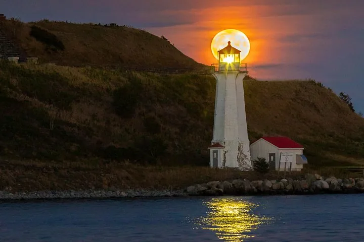Grande pleine lune se couchant juste derrière un phare blanc au crépuscule